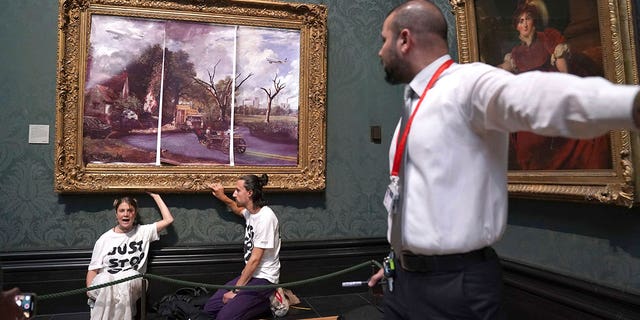 A security guard looks at protesters who glued their hands to the frame of John Constable's The Hay Wain, inside the National Gallery, London, Monday July 4, 2022. Police say two climate change protesters have been arrested after they glued themselves to the frame of a famous John Constable painting hanging in Britain’s National Gallery. The two, from the protest group "Just Stop Oil," stepped over a rope barrier and covered "The Hay Wain" on Monday with large sheets of paper depicting "an apocalyptic vision of the future" of the landscape. 