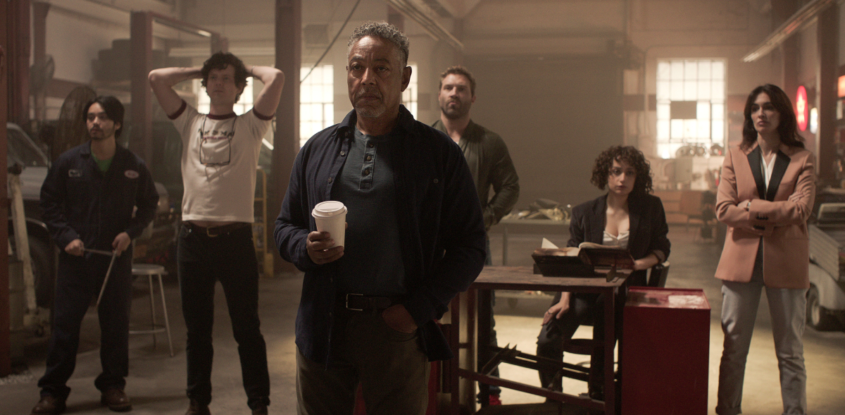 Leo Pap and his crew in the Netflix series Kaleidoscope stand around in what appears to be a warehouse. Leo is holding a coffee cup.
