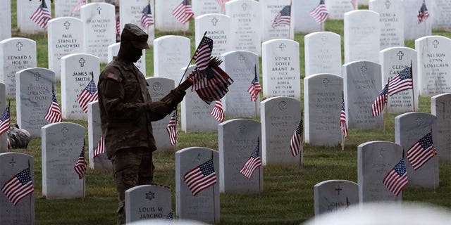 Military member holding flags and placing them on a tombstone.