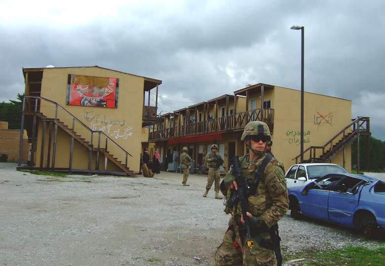 A soldier stands in front of a mock Afghan village, holding his rifle.
