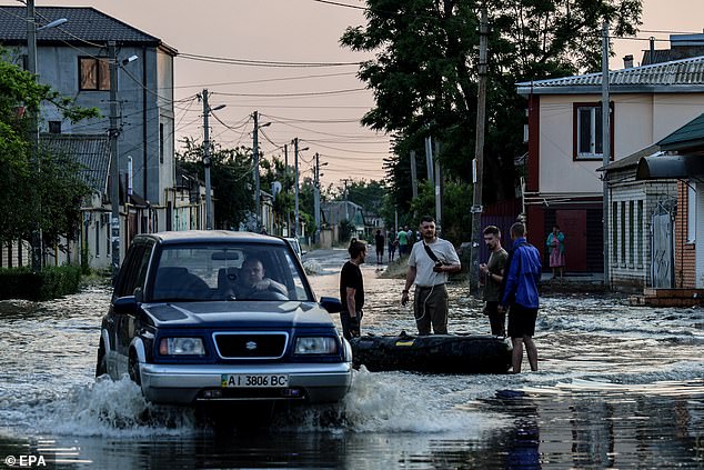 A car makes its way past people stanidng next to an inflatable boat in a flooded street of Kherson, Ukraineon Tuesday