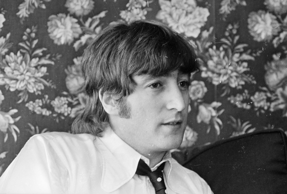 A black and white picture of John Lennon sitting in front of floral wallpaper. 