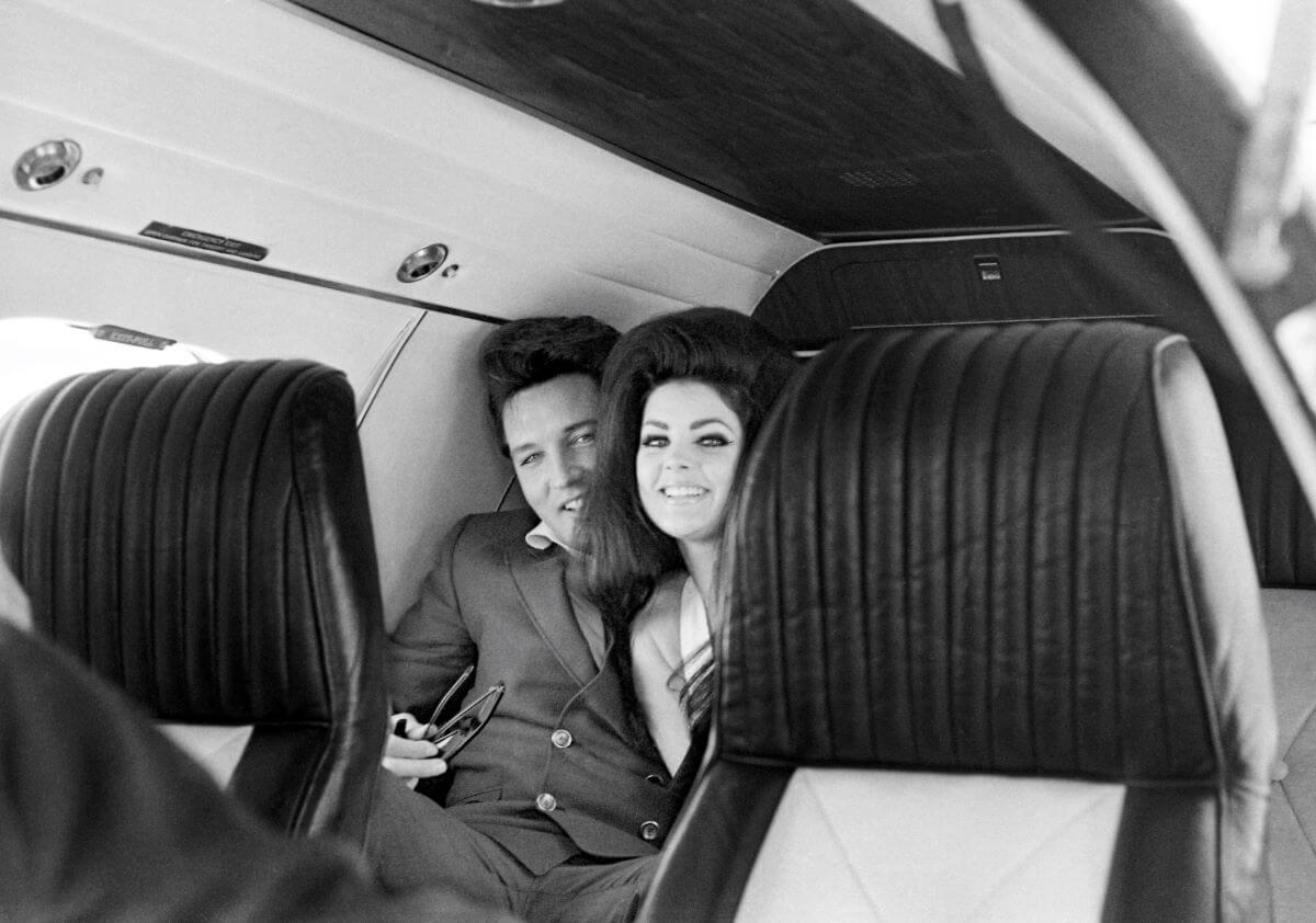 A black and white picture of Elvis and Priscilla Presley sitting on a plane together. They look at the camera from between two seats.