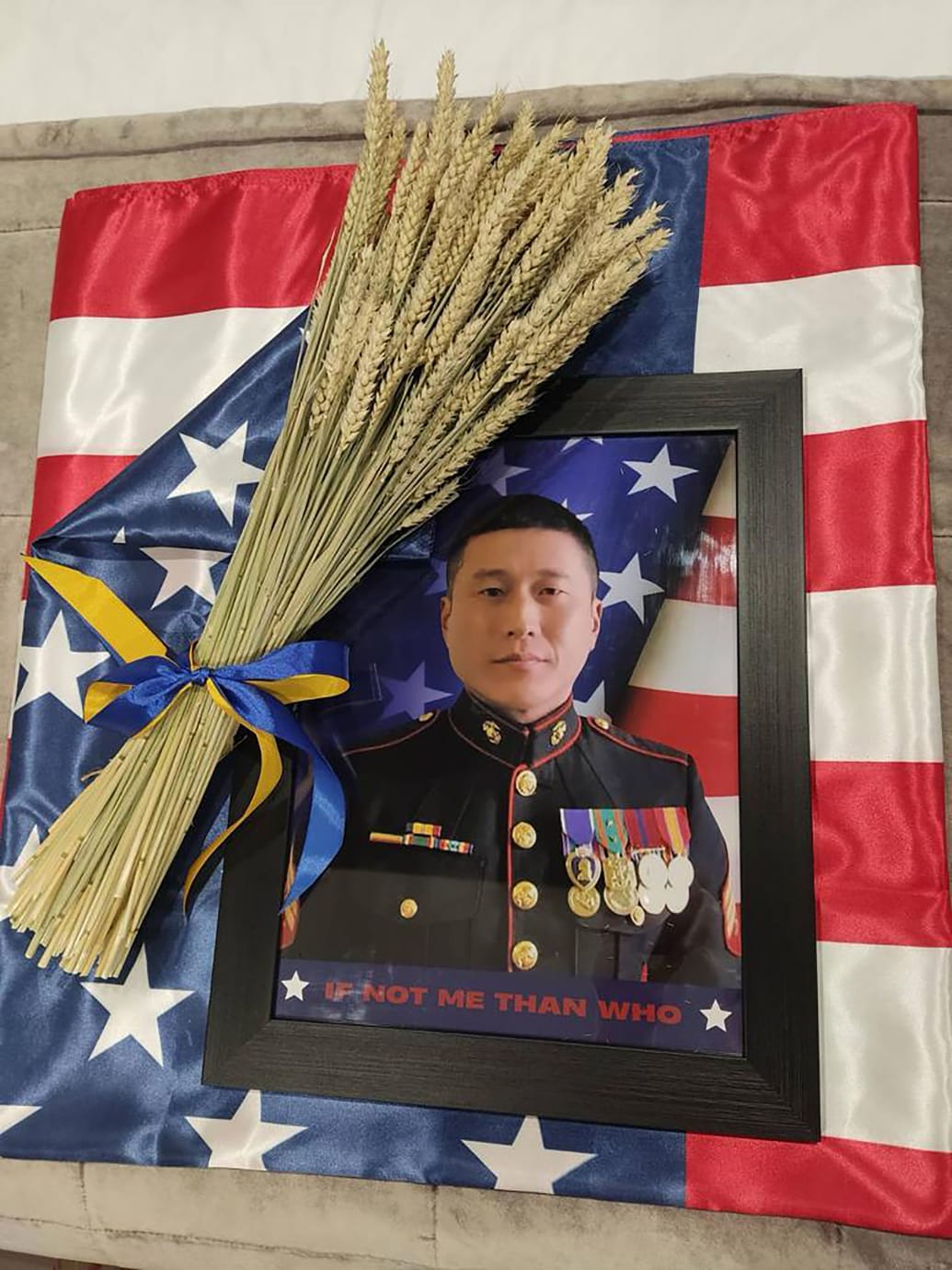 A picture shows a photograph of Grady Kurpasi in his military uniform laid on top of an American flag. A bundle of wheat tied together in Ukrainian blue and yellow lays on top. 